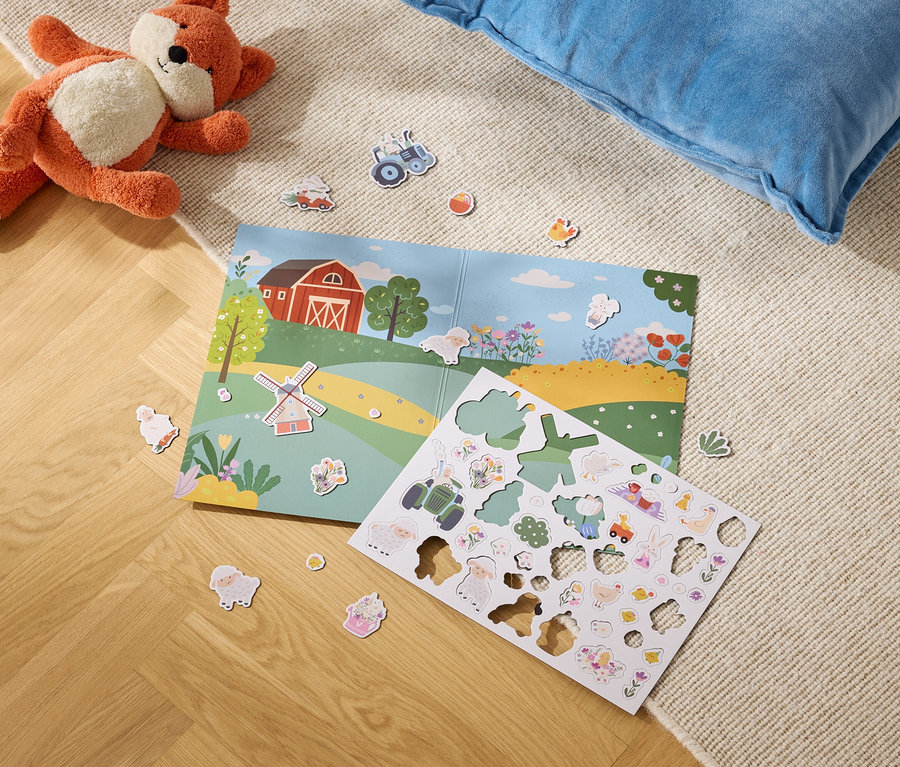 Plyšová líška leží vedľa otvorenej knihy s ilustráciou farmy a hárku s nálepkami s farmou na koberci.