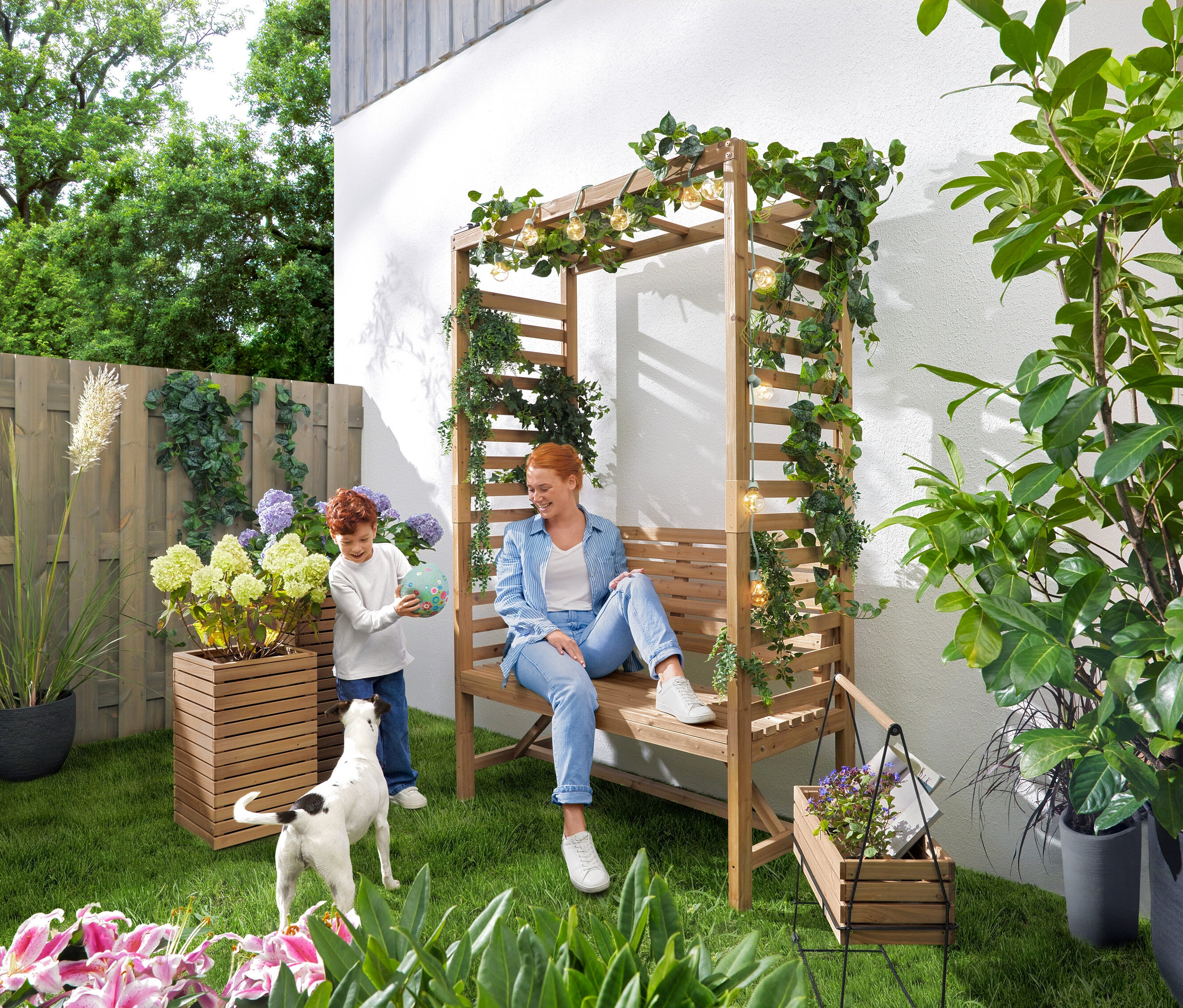 Žena sedí na záhradnej lavičke s pergolou, zatiaľ čo sa dieťa hrá s loptou. Nad lavicou visí LED svetelná reťaz »Glühbirnen«. V záhrade stoja stĺpovité kvetináče »Tinus«, 2 ks a krabička na bylinky s podstavcom »Elin«.