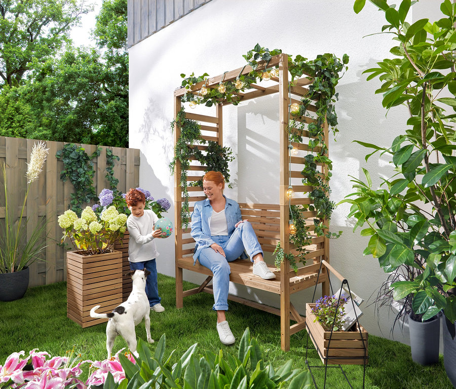 Žena sedí na záhradnej lavičke s pergolou, zatiaľ čo sa dieťa hrá s loptou. Nad lavicou visí LED svetelná reťaz »Glühbirnen«. V záhrade stoja stĺpovité kvetináče »Tinus«, 2 ks a krabička na bylinky s podstavcom »Elin«.