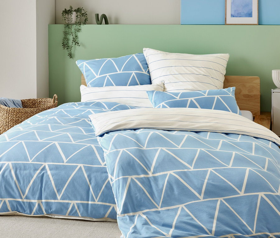 Posteľ s bledomodrým poťahom s bielymi trojuholníkmi a pruhovanými obliečkami na vankúše, vedľa prúteného koša a nočného stolíka s lampou.