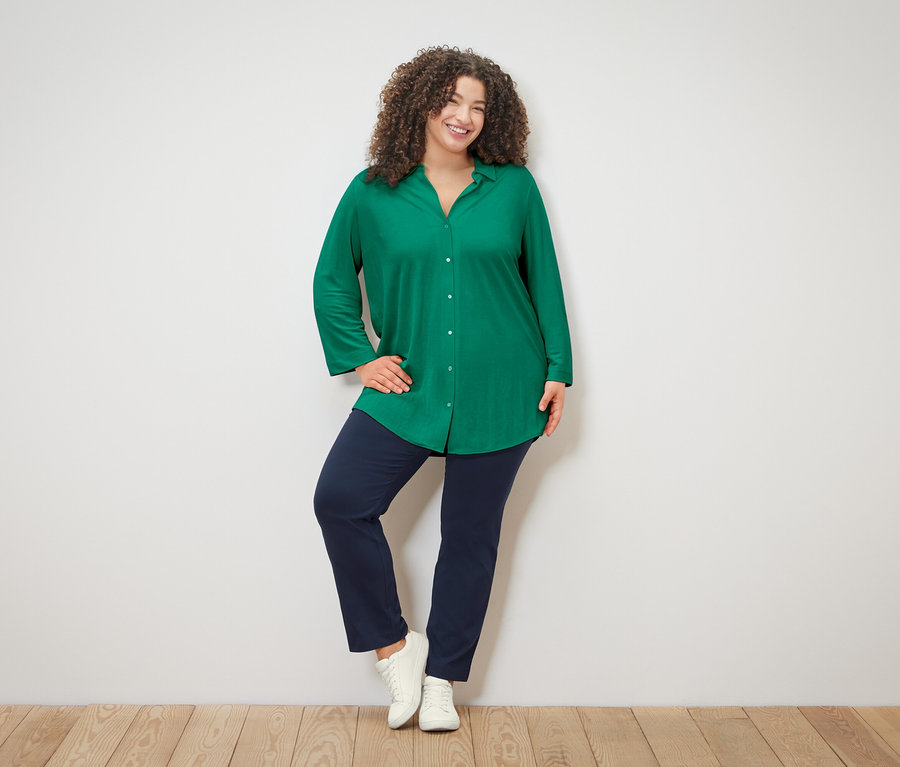 Žena s kučeravými vlasmi v zelenej blúzke, modrých nohaviciach a bielych teniskách pózuje na bielom pozadí.