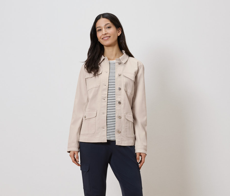 Žena má na sebe béžovú bundu cez pásikavú košeľu a tmavomodré nohavice.