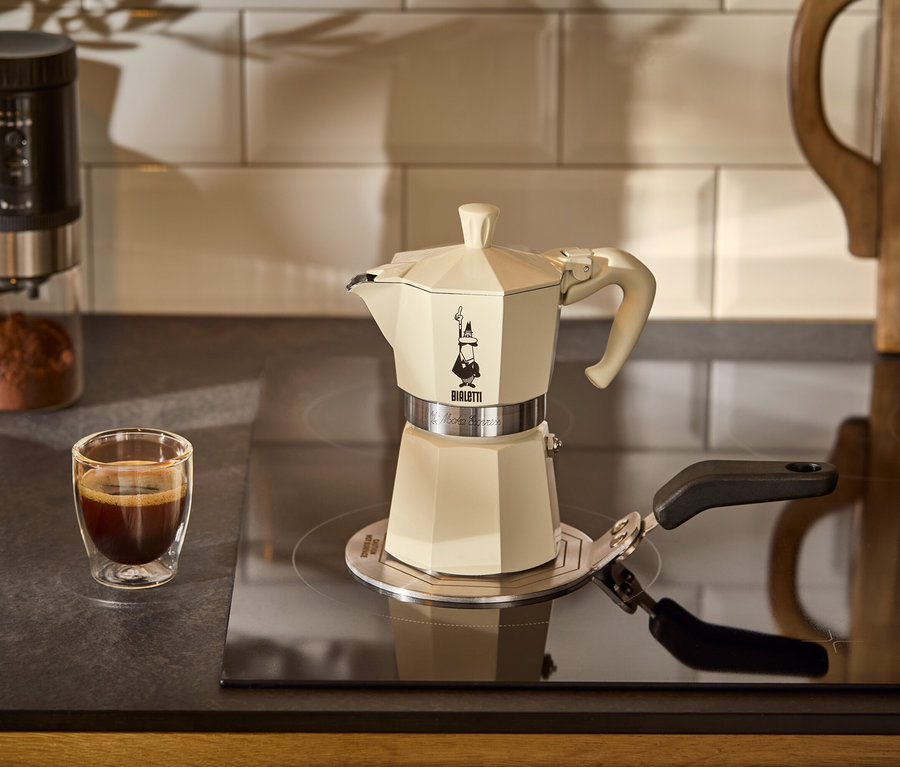 Krémový kávovar Bialetti Moka Express, 190 ml stojí na adaptérové podložke na indukciu Bialetti na varnej doske. V pozadí je šálka kávy.