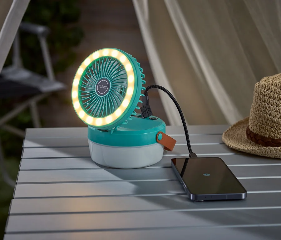 Tyrkysový kempingový ventilátor so svetelným kruhom nabíja smartfón na kempingovom stole, vedľa neho je slamený klobúk.