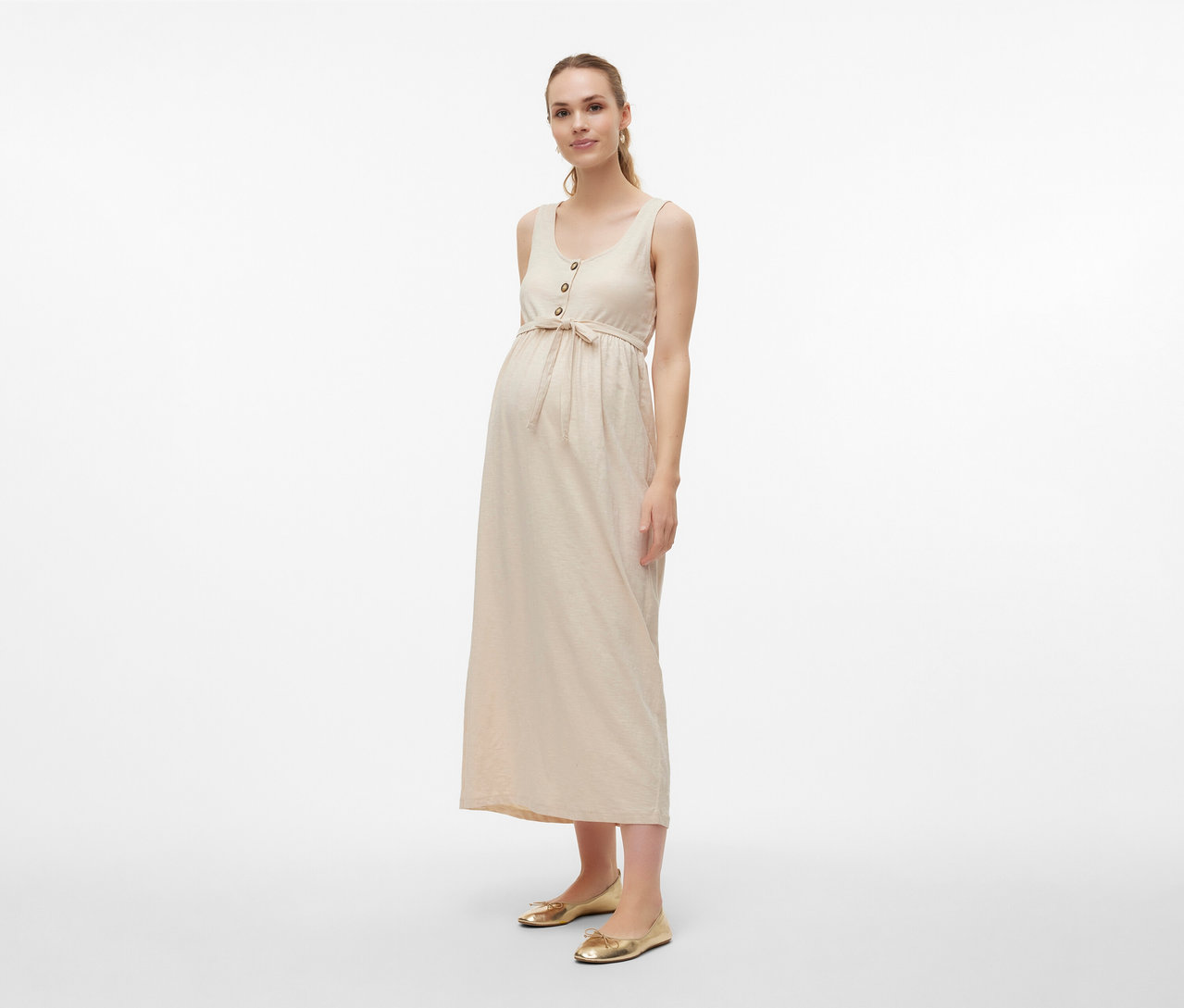 Žena nosí tehotenské šaty MAMALICIOUS »MLEVI LIA« s opaskom a zlaté baleríny na svetlom pozadí.