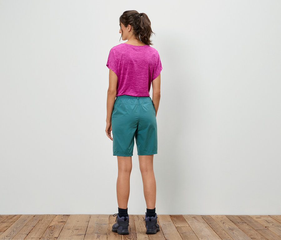 Žena stojí chrbtom k fotoaparátu, má na sebe ružové tričko a zelené šortky.