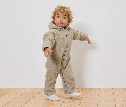 Malé dieťa s kučeravými blond vlasmi má na sebe béžový teddy overal s kapucňou a biele ponožky, stojí na drevenej podlahe.