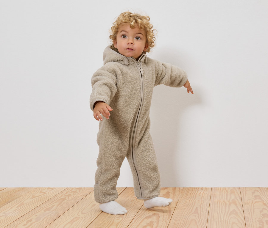 Malé dieťa s kučeravými blond vlasmi má na sebe béžový teddy overal s kapucňou a biele ponožky, stojí na drevenej podlahe.