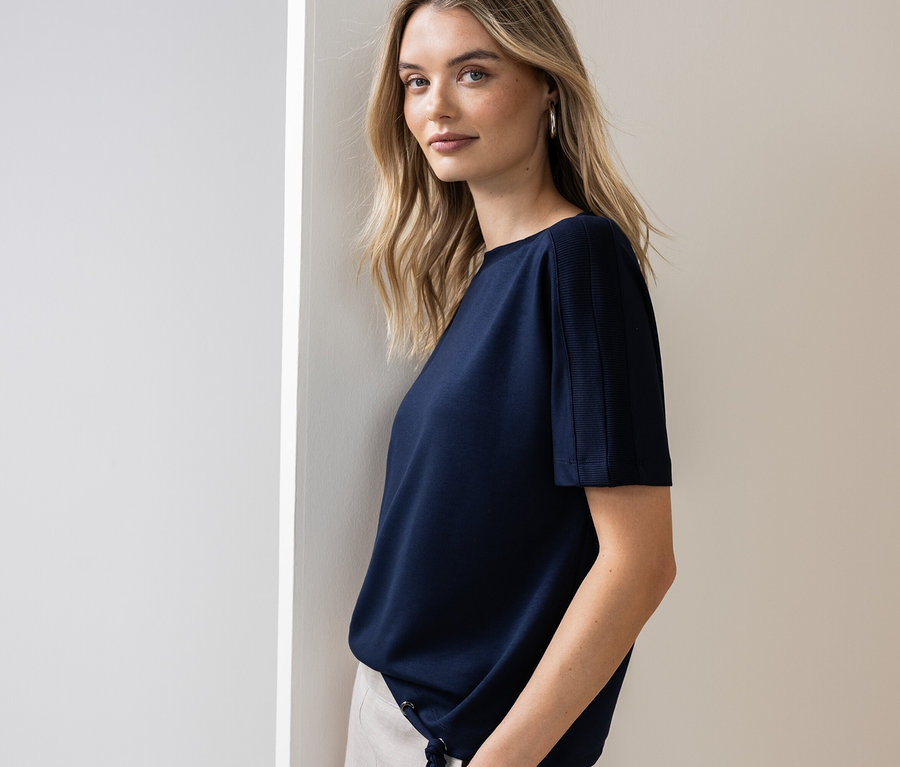 Žena sa opiera o stenu a má na sebe tmavomodré tričko a svetlé nohavice.