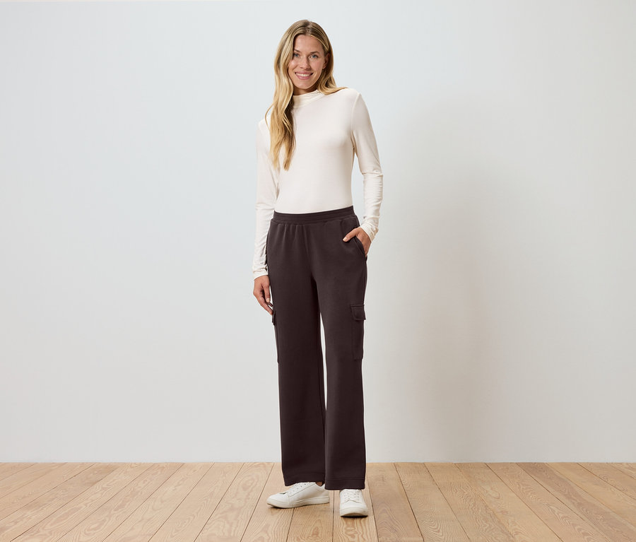 Žena stojí s rukou vo vrecku pred stenou a má na sebe hnedé cargo nohavice.