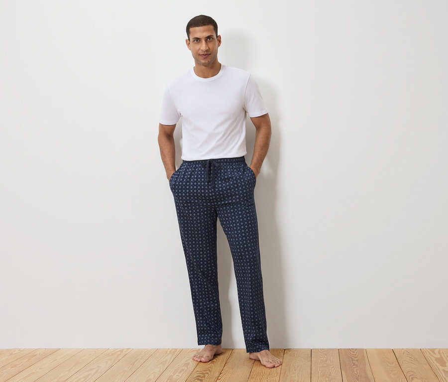 Muž stojí pred bielou stenou, má na sebe biele tričko a tmavomodré pyžamové nohavice s bodkovaným vzorom.