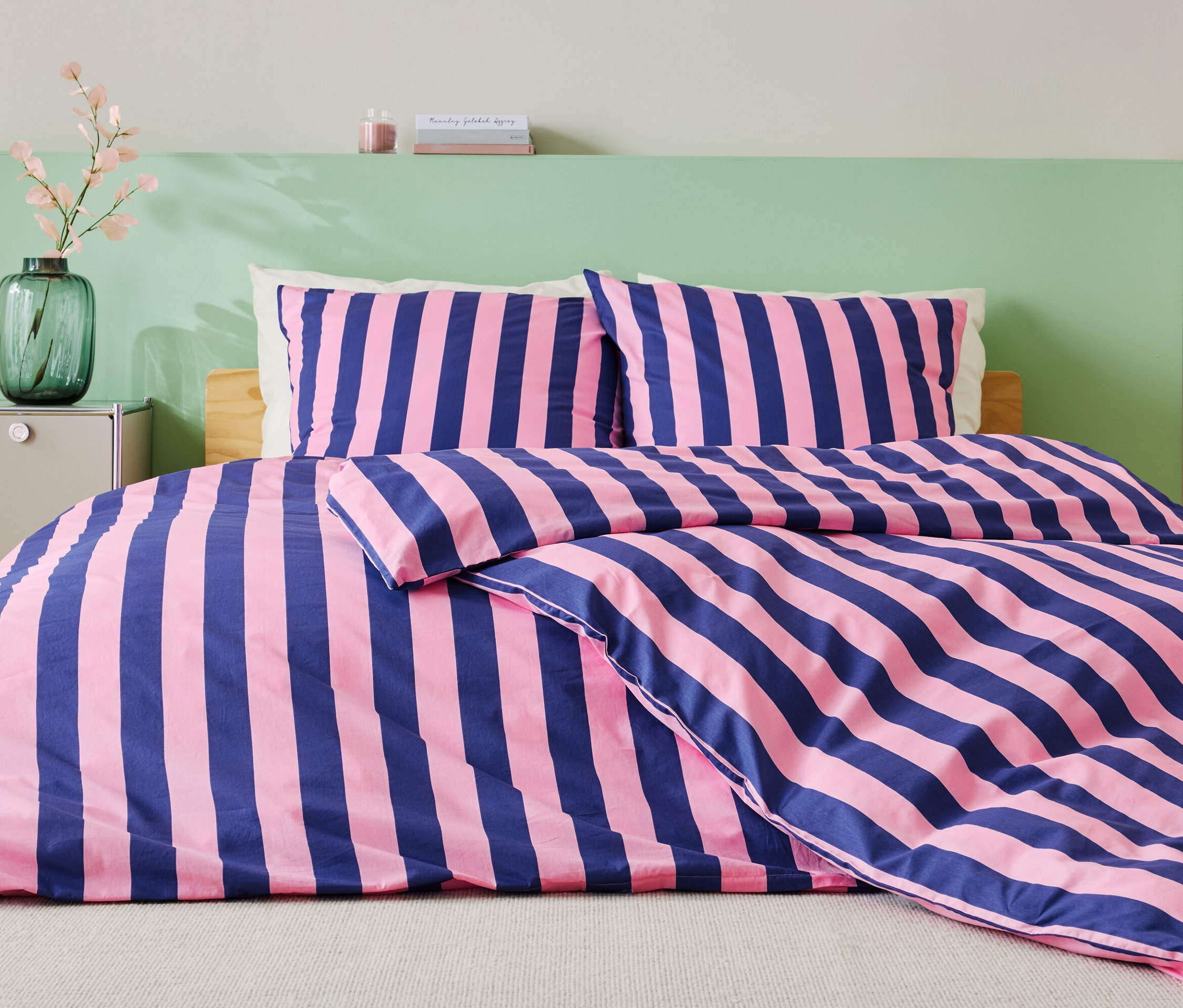 Posteľ s modro-ružovými pruhovanými obliečkami, dvoma vankúšmi a zelenou stenou v pozadí.