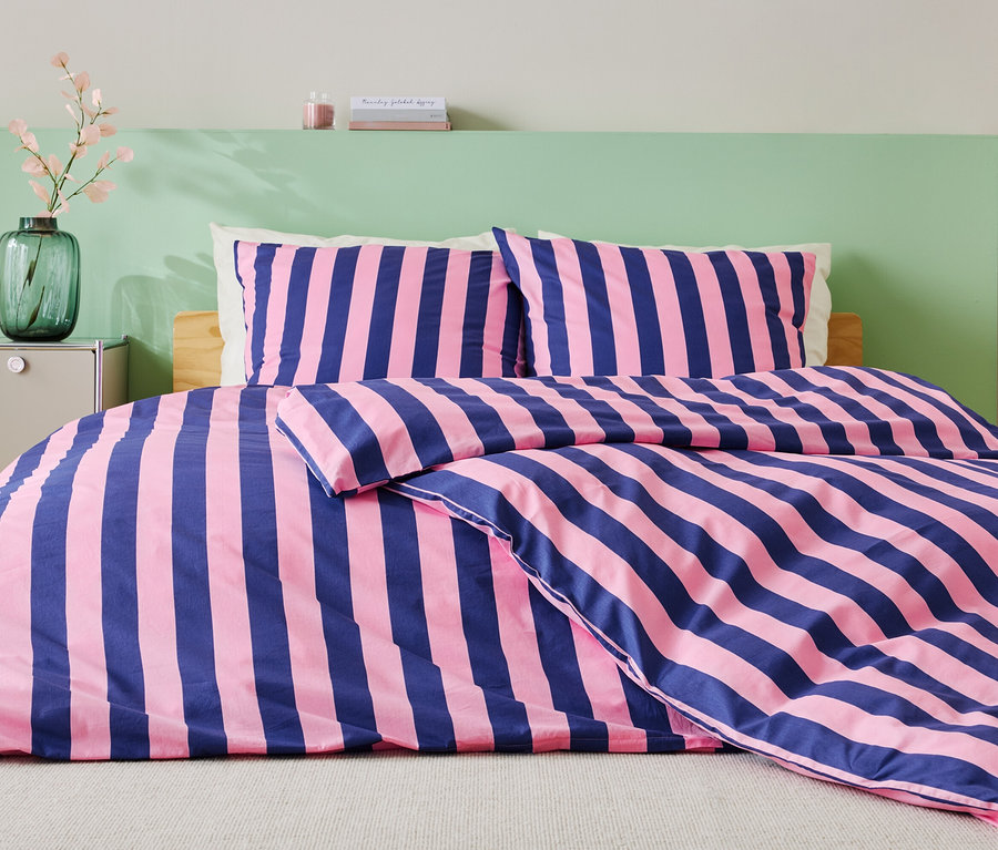 Posteľ s modro-ružovými pruhovanými obliečkami, dvoma vankúšmi a zelenou stenou v pozadí.