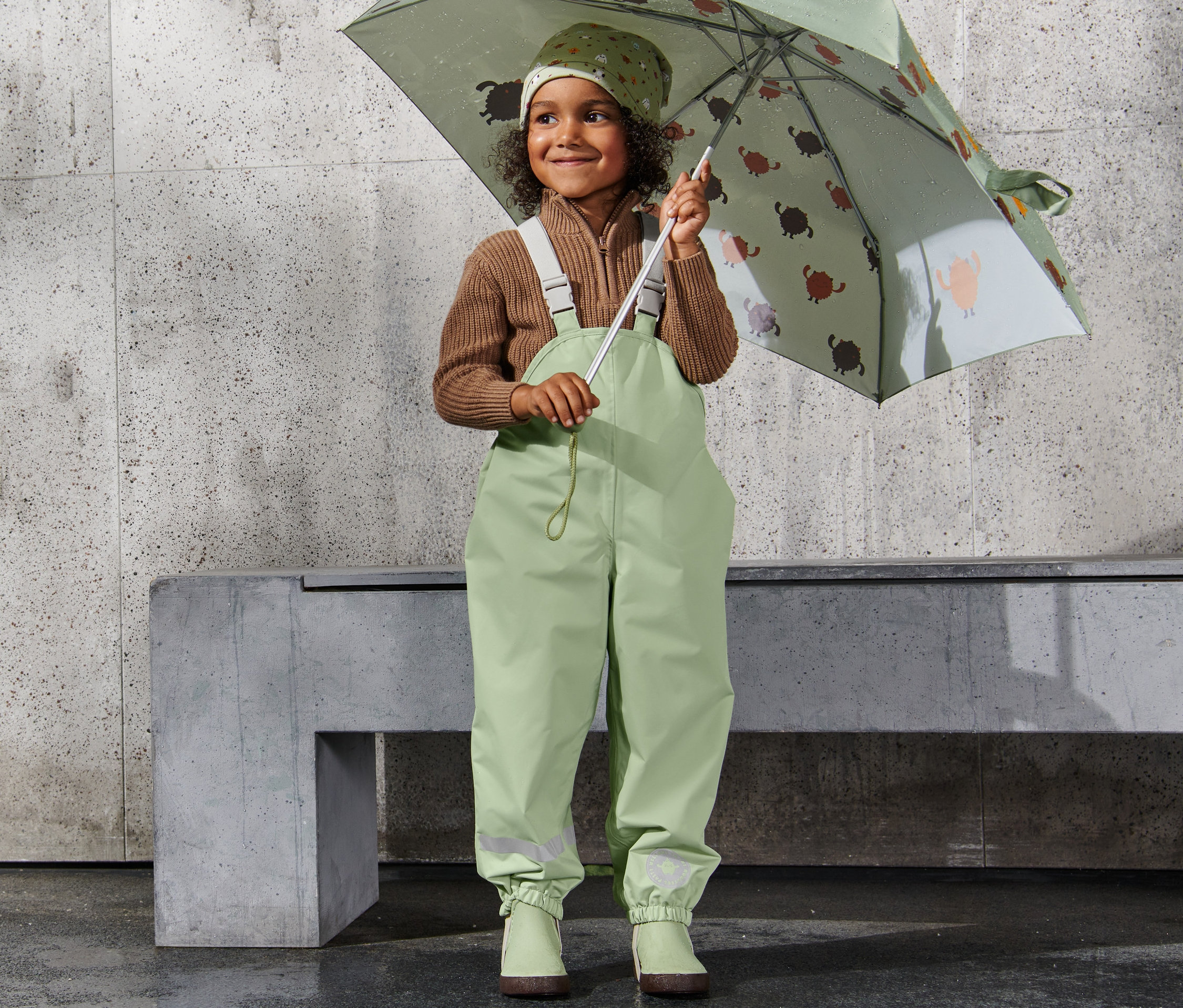 Usmievajúce sa dievčatko drží dáždnik s potlačou a má na sebe Nohavice do dažďa.