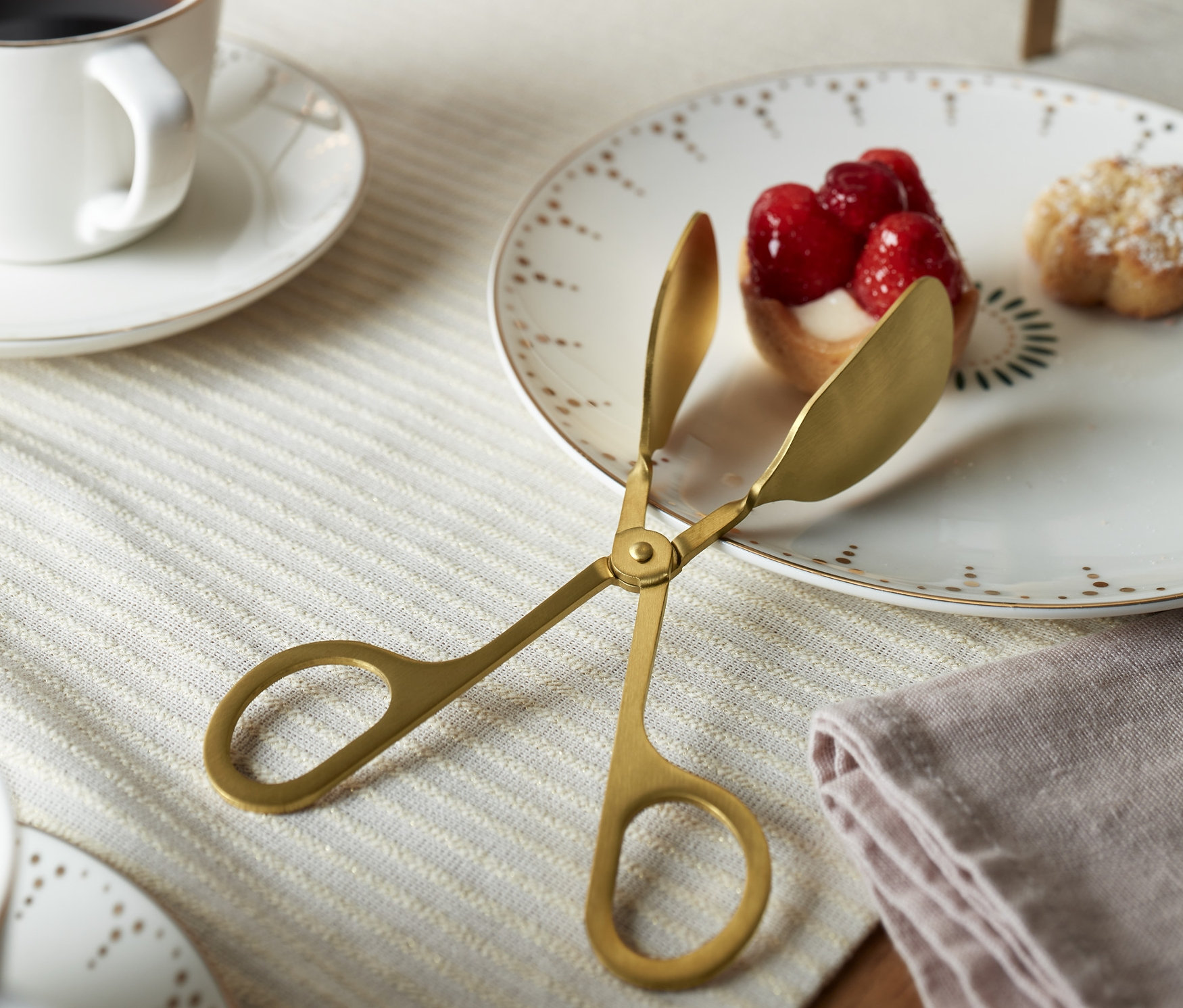 Zlaté kliešte na pečivo ležia na tanieri vedľa mini koláčikov s jahodami.