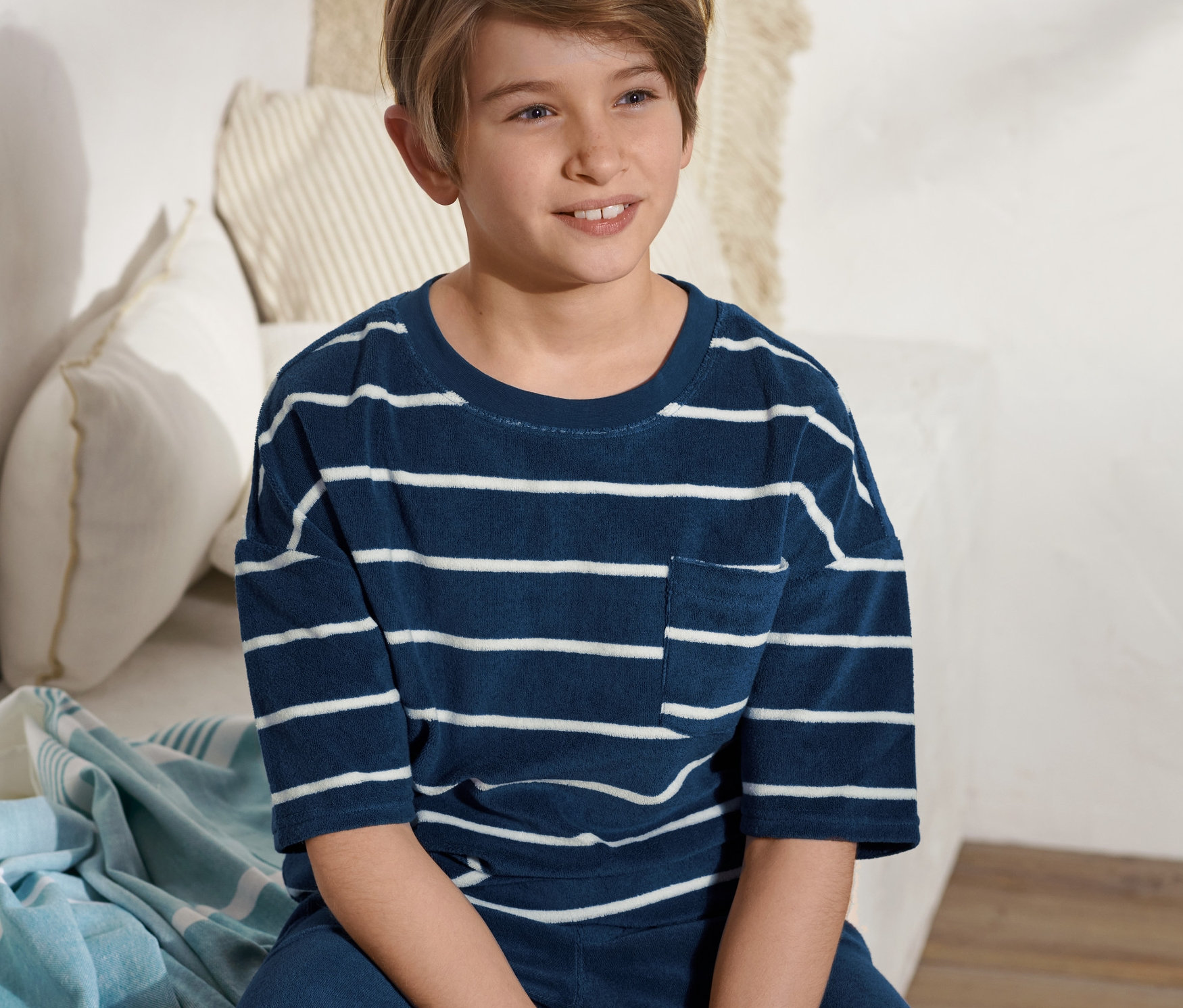 Chlapec sedí na posteli a má na sebe froté tričko s modrými a bielymi pruhmi.