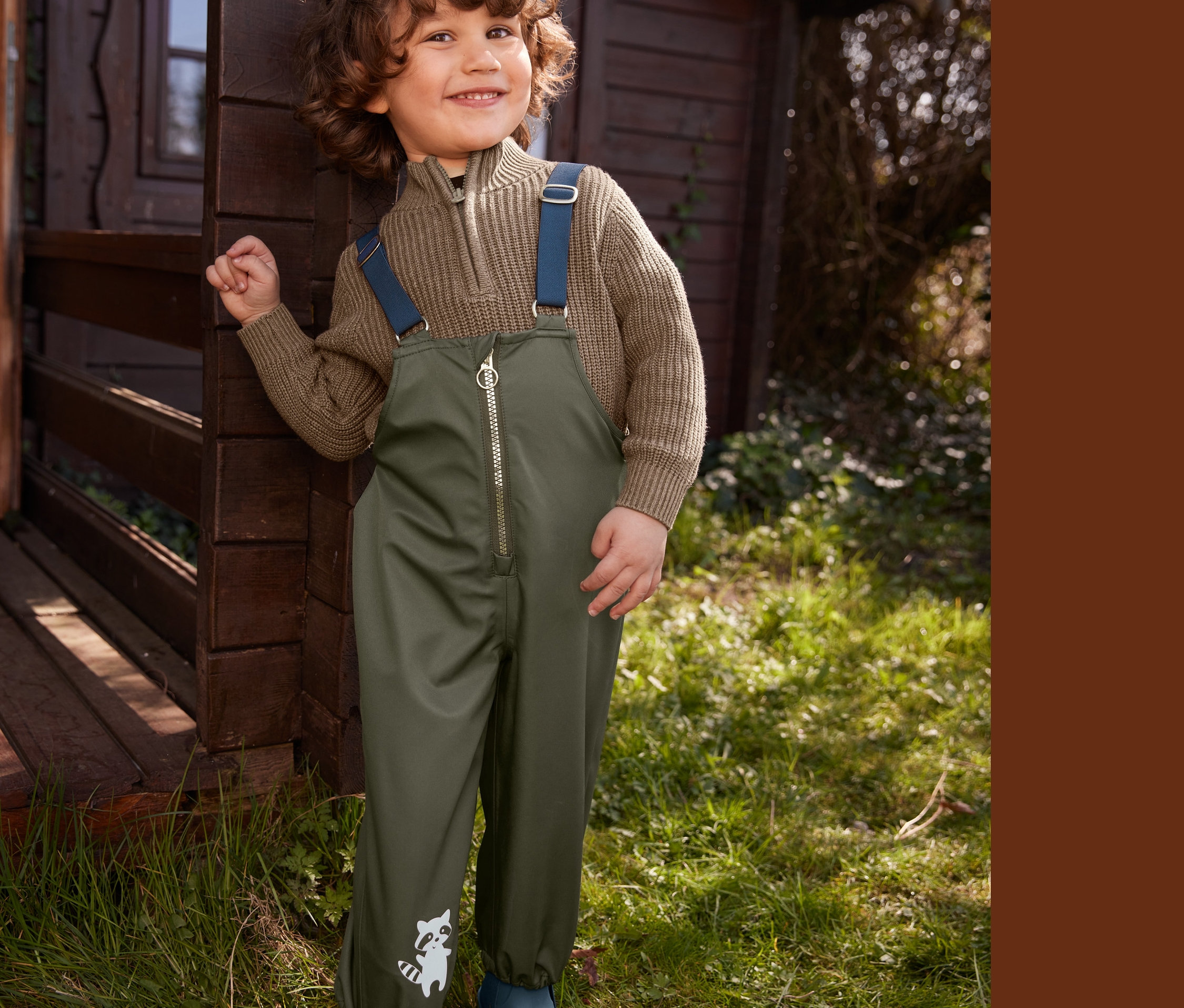 Chlapec stojí pri drevenej búde a má na sebe hnedý pulóver, nohavice do dažďa a gumové čižmy.