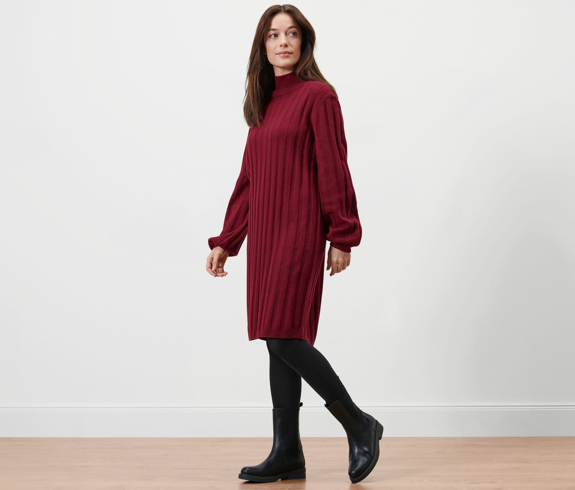 Žena kráča a má na sebe rebrované pletené šaty so stojačikom, čierne legíny a topánky.