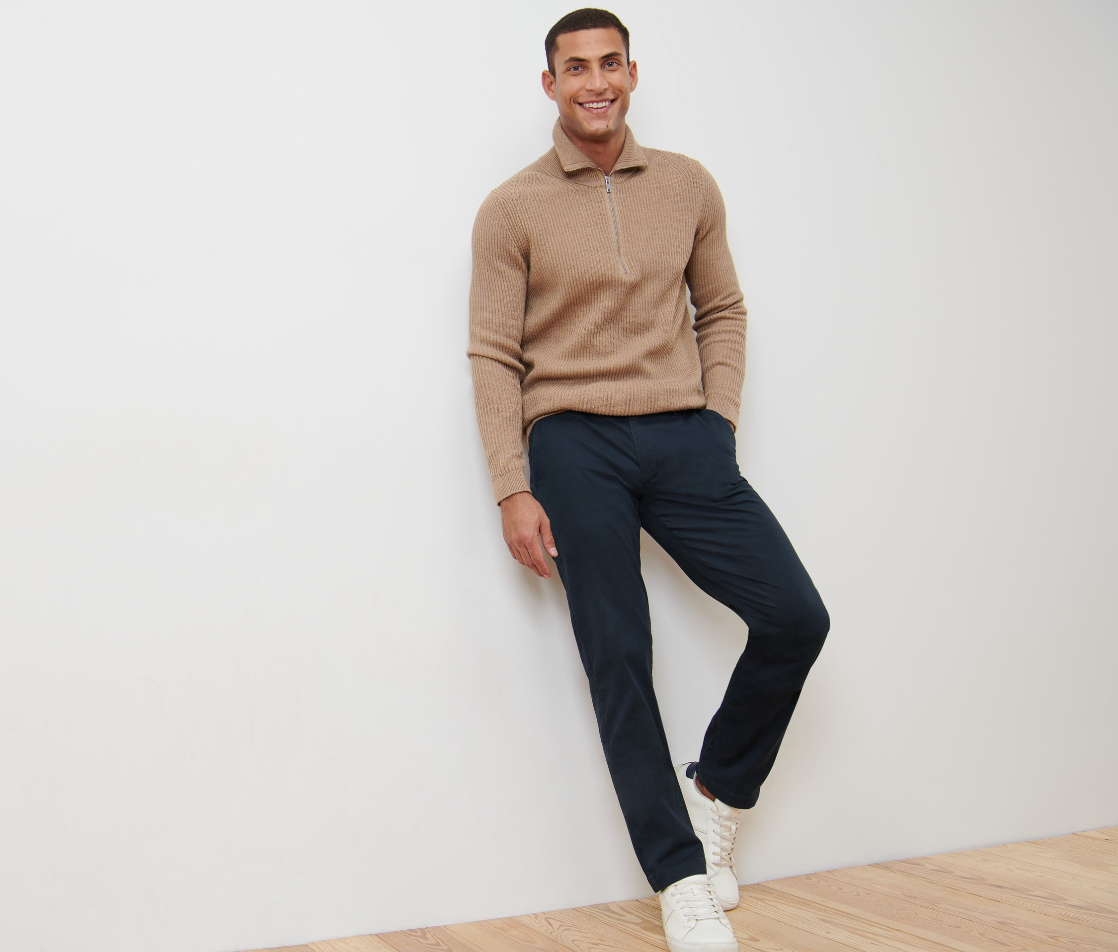 Muž sa usmieva a opiera sa o stenu v námorníckych modrých chino nohaviciach.