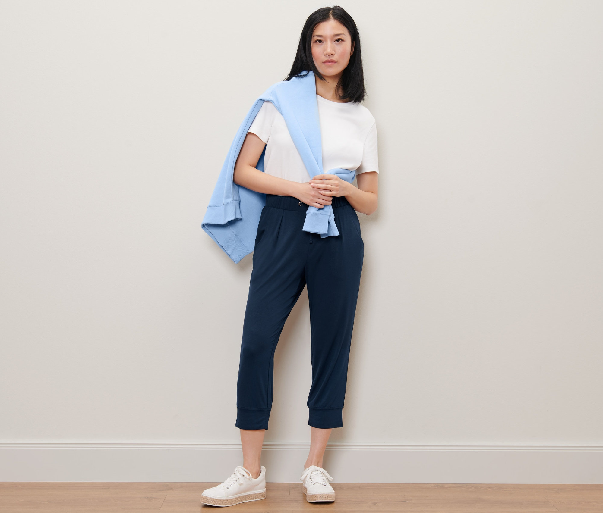 Žena stojí a má na sebe 3/4 džersejové nohavice, biele tričko a cez ramená prehodený modrý sveter.