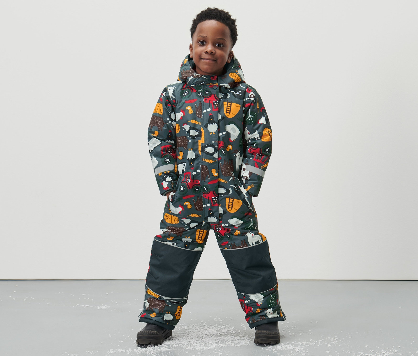 Chlapec v detskom zimnom overale s celoplošnou potlačou s motívom gazdovského dvora stojí na podlahe.