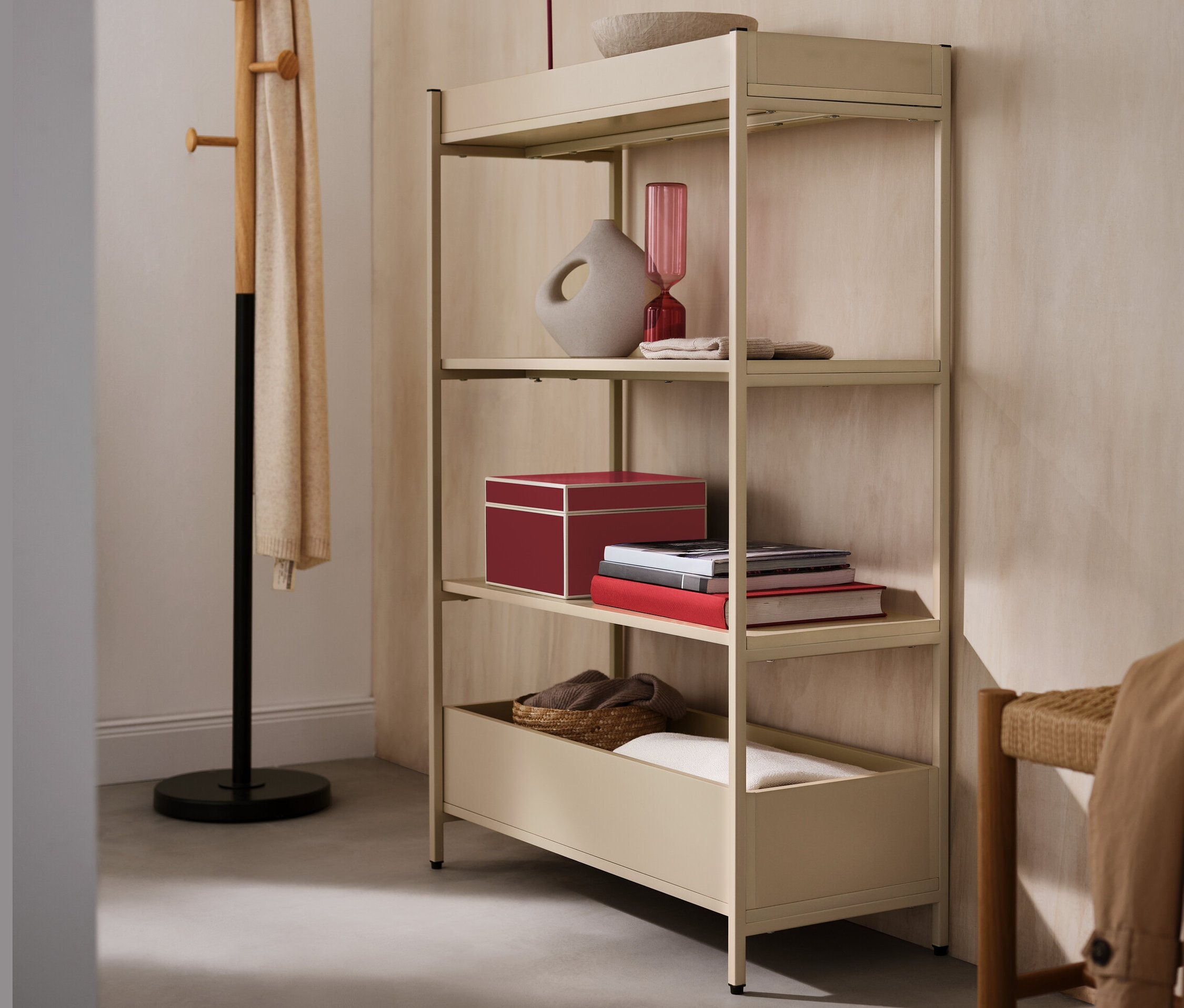 Béžová konzolová skrinka so štyrmi policami, na ktorej je červená stolová LED lampa s akumulátorom, váza a miska. Vedľa stojí stojan na oblečenie.