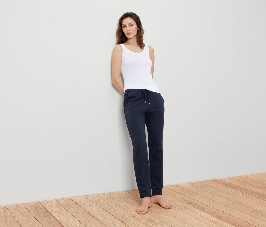 Žena opretá o stenu má na sebe biele tielko a tmavomodré tepláky.