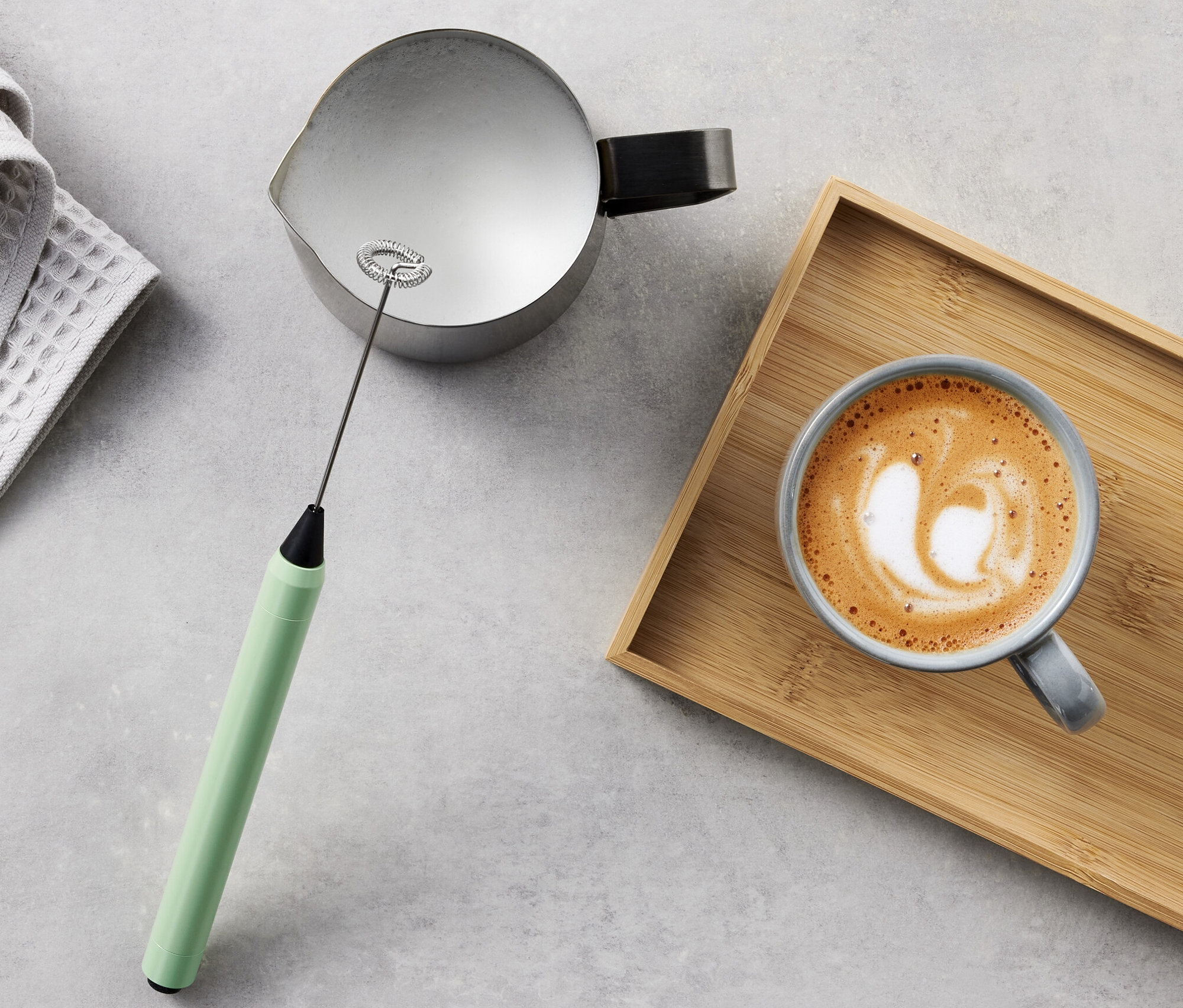 Sivá utierka, nádoba, napeňovač mlieka a káva s mliekom na drevenom podnose.