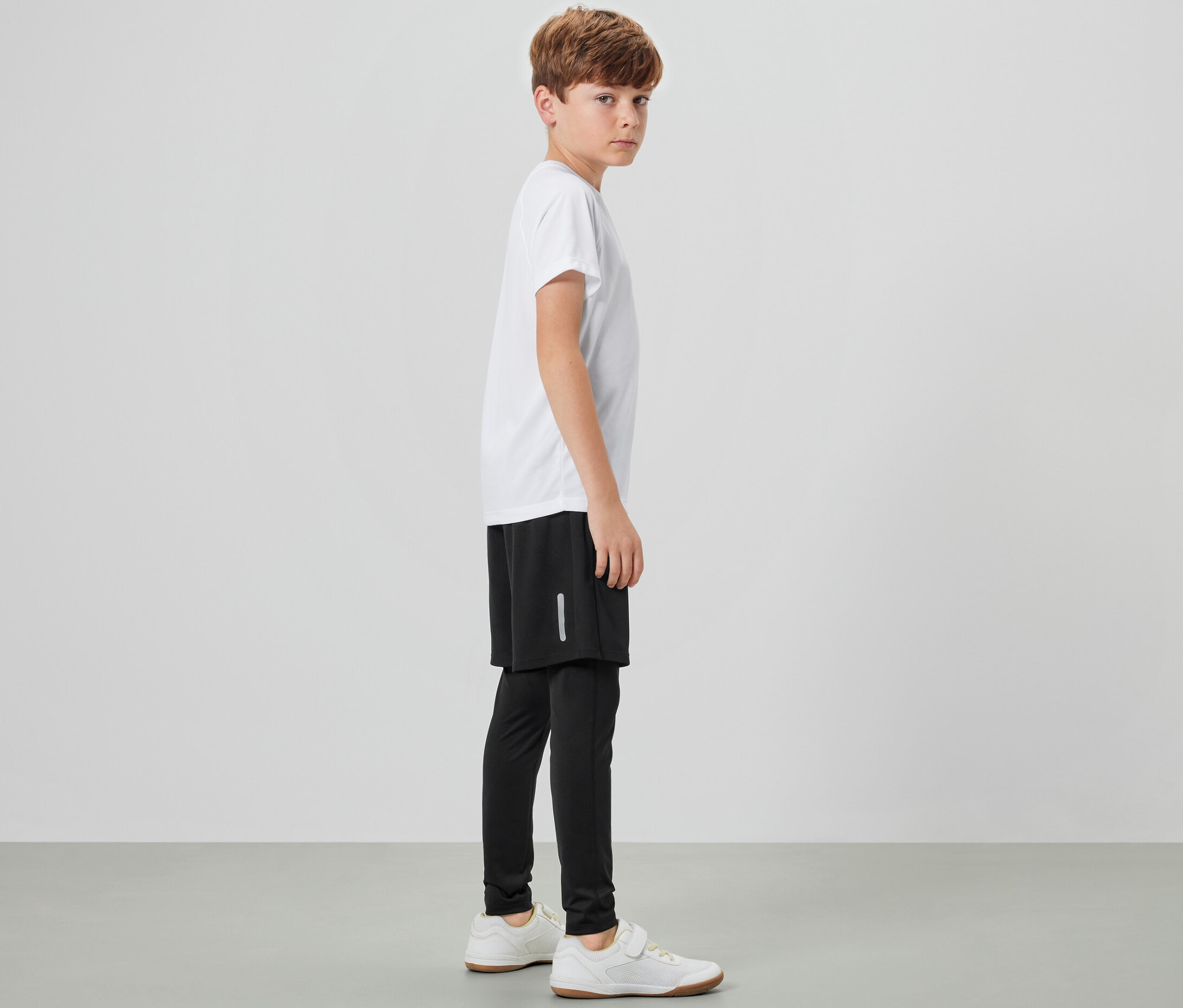 Chlapec stojí bokom a má na sebe detské športové legíny so šortkami, biele tričko a tenisky.