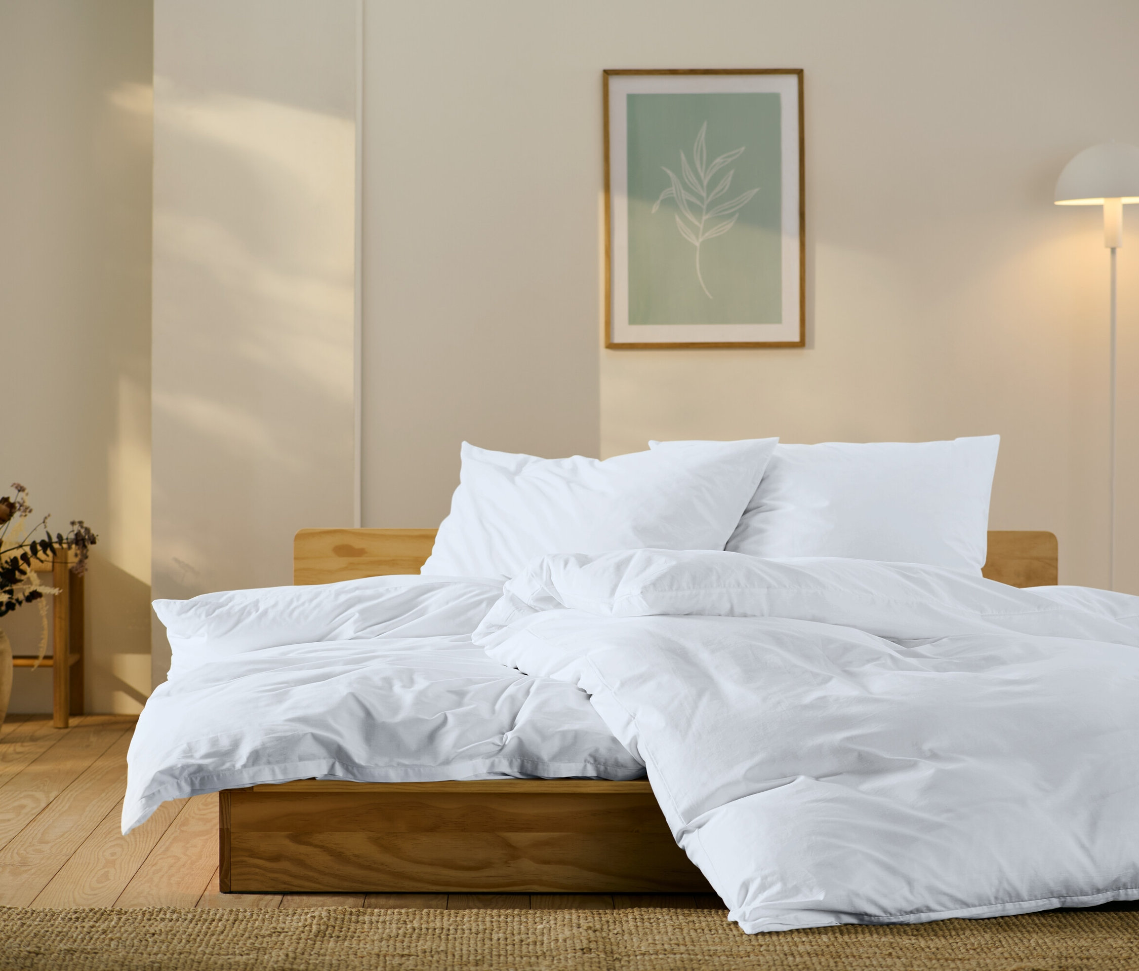 Drevená posteľ s bielymi obliečkami, vankúšmi a prikrývkou. Na stene obraz s motívom listu a lampa.