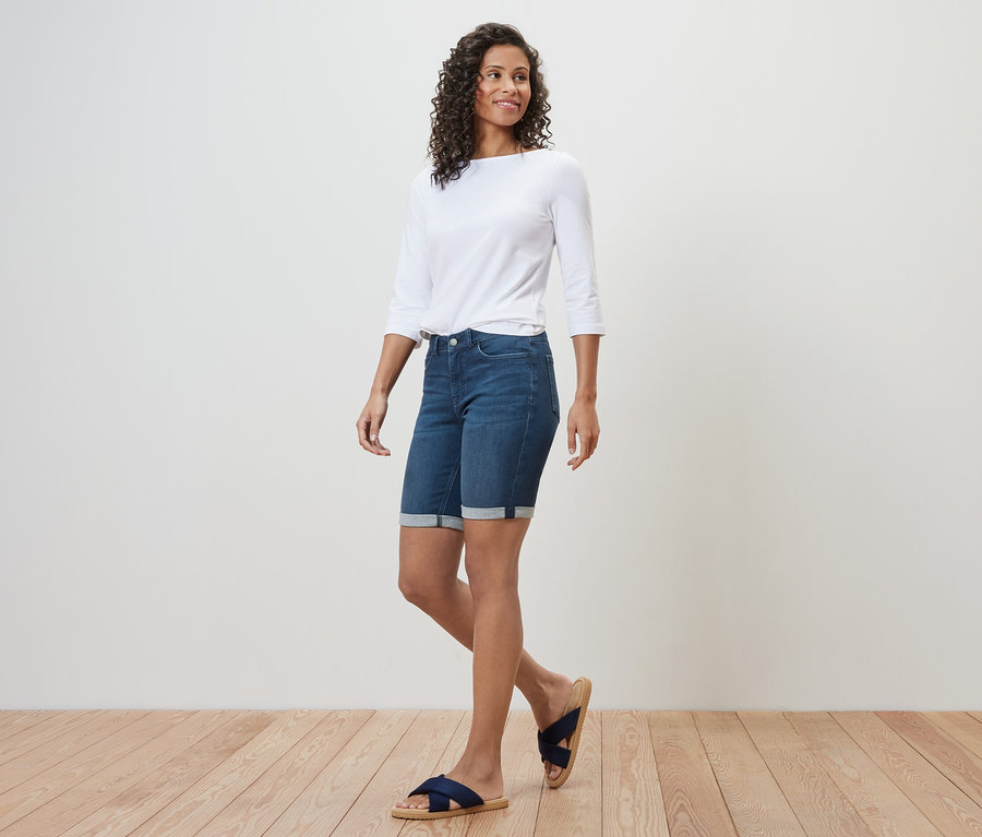 Žena kráča a má na sebe džínsové bermudy – strih »Lea«, tmavomodrý denim, biely top s trojštvrťovými rukávmi a sandále.