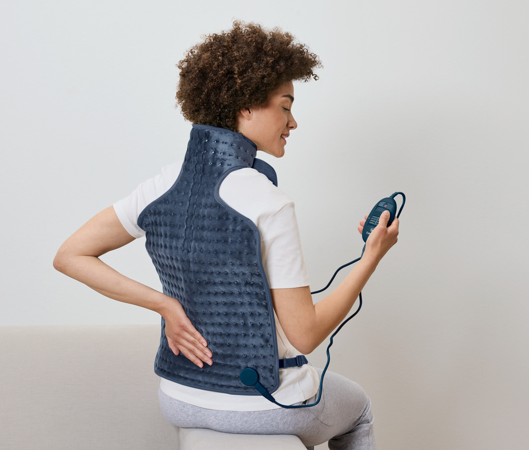 Žena sedí a používa vyhrievaciu dečku na chrbát s ovládačom v ruke.