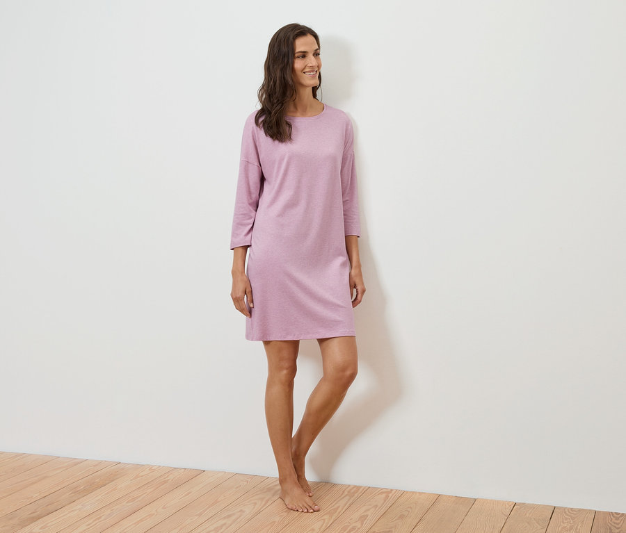 Žena stojí a usmieva sa, má na sebe fialové šaty s trojštvrťovými rukávmi.