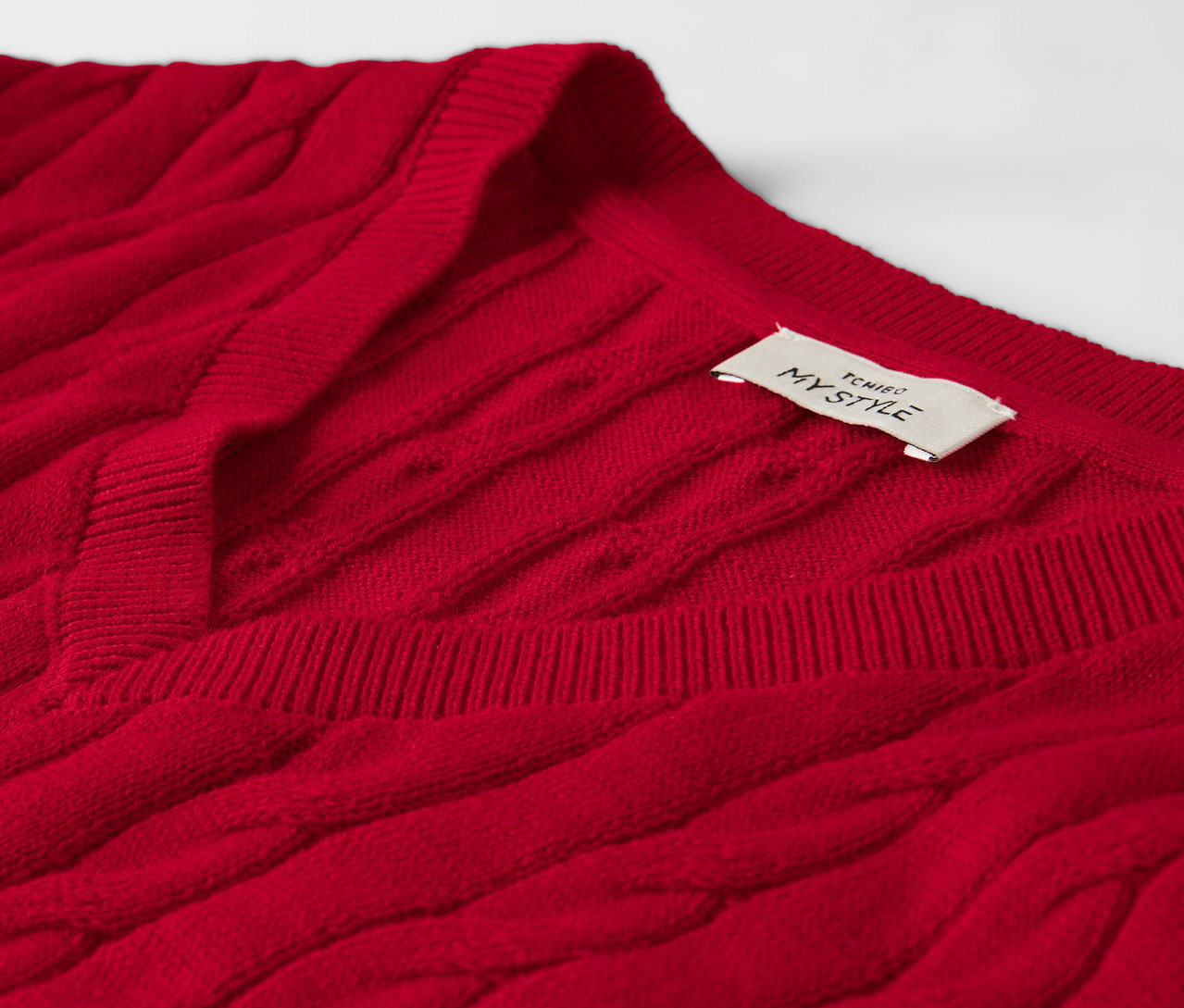 Detail červeného svetra s vrkočovým vzorom a štítkom pri krku.