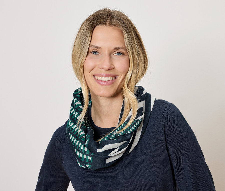 Žena s blond vlasmi a úsmevom má na sebe tmavomodrý sveter a šál s geometrickým vzorom v zelenej a bielej farbe.