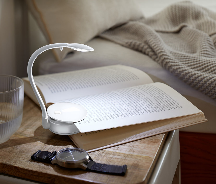 Na nočnom stolíku leží otvorená kniha osvetlená malou lampičkou, vedľa nej stojí pohár s vodou a hodinky.