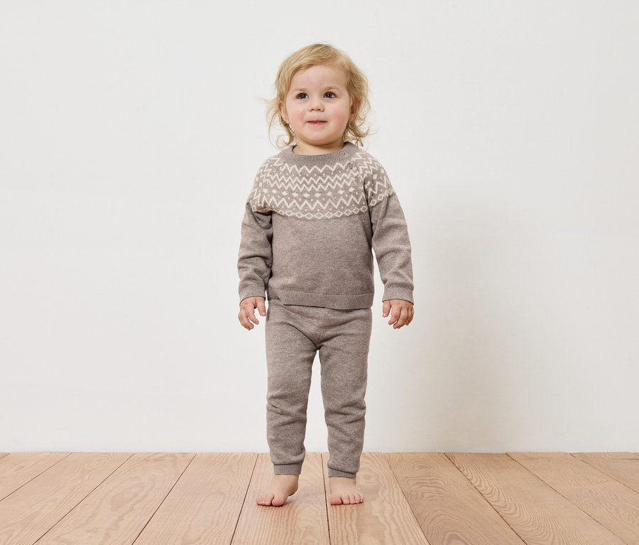 Batoľa stojí na drevenej podlahe a má na sebe hnedý sveter a nohavice.