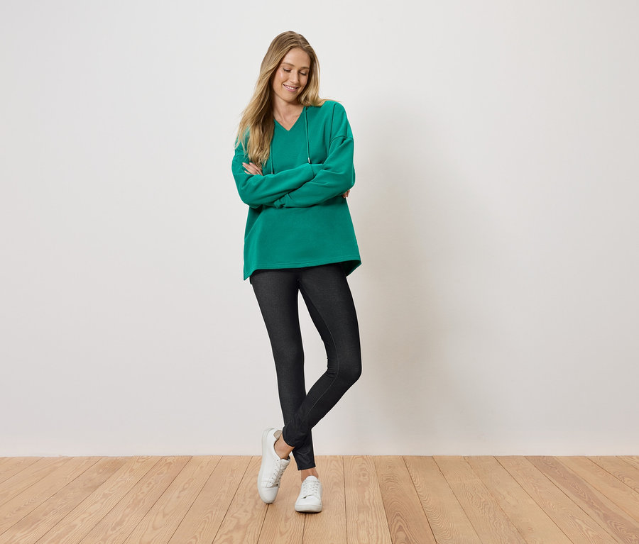 Žena stojí s prekríženými rukami a usmieva sa, oblečená do zeleného svetra, čiernych legín a bielych topánok.