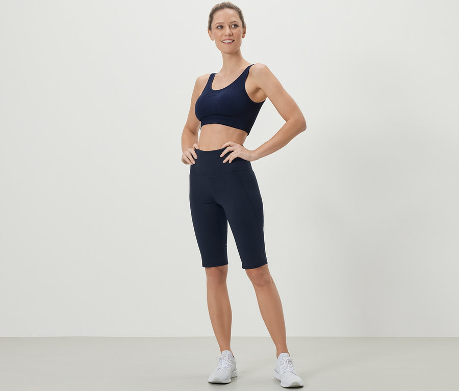 Žena pózuje v tmavomodrej športovej podprsenke a športových cyklistických šortkách.