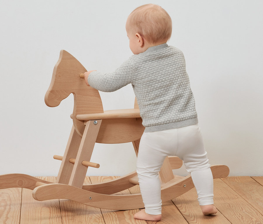Batoľa v detskom pletenom svetríku sa opiera o dreveného hojdacieho koňa.