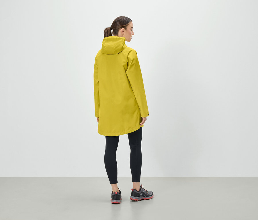 Žena v žltej bunde s kapucňou, čiernych legínach a športovej obuvi stojí chrbtom k fotoaparátu.