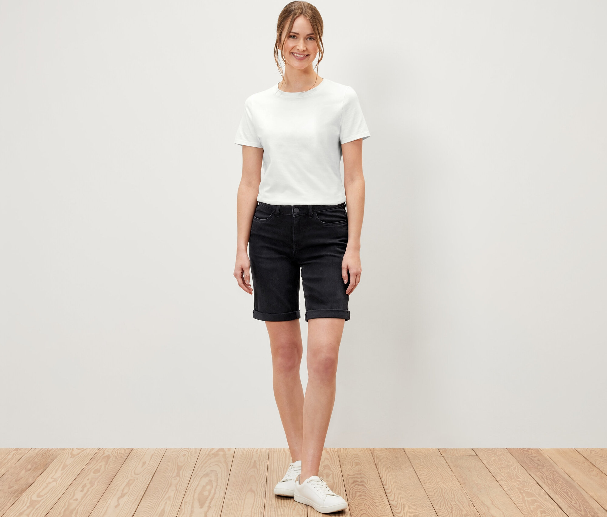Žena stojí na drevenej podlahe a má na sebe džínsové bermudy – strih »Lea«, čierny denim a biele tričko.
