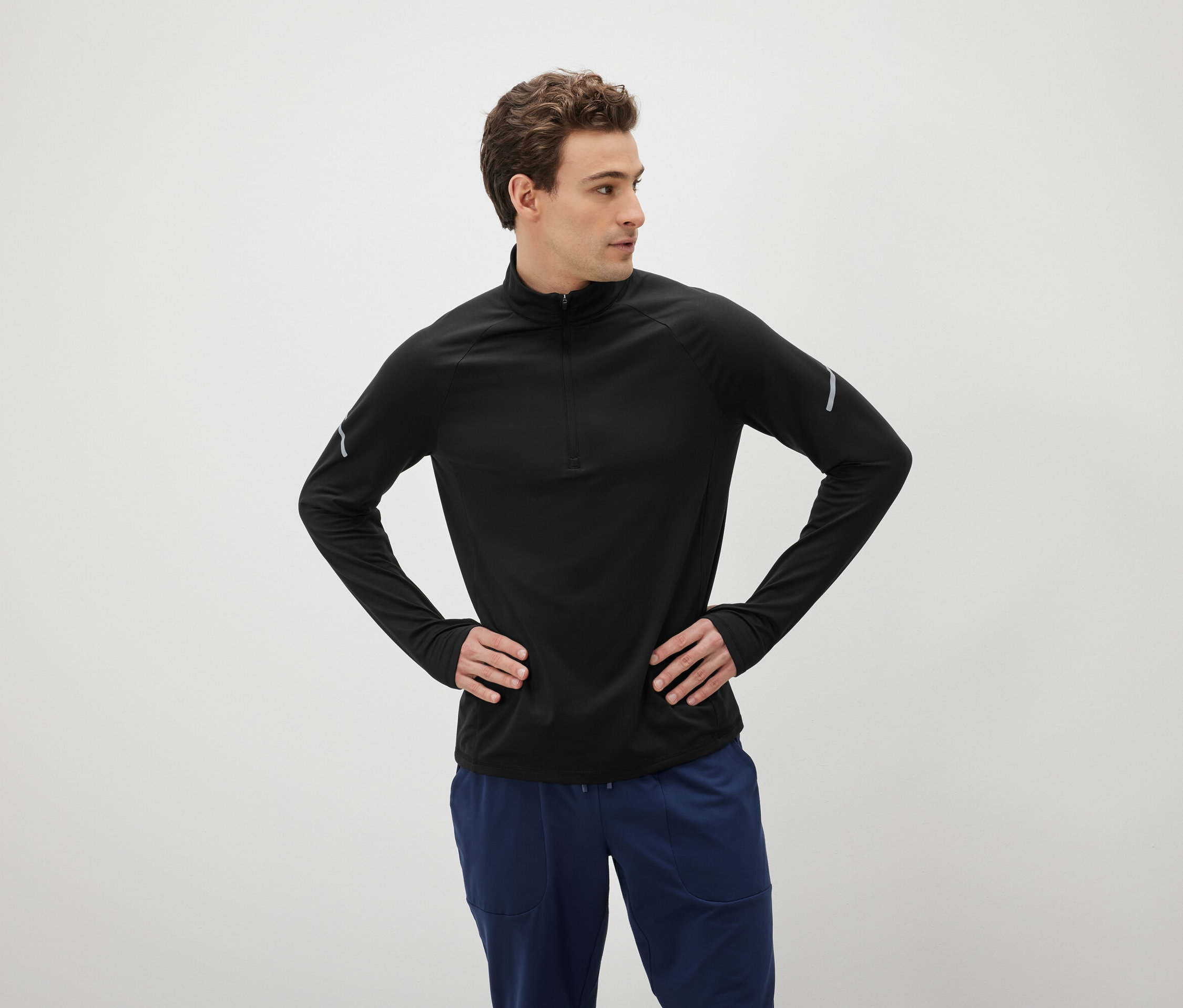 Muž s hnedými vlasmi pózuje s rukami vbok oblečený v čiernom tričku s dlhým rukávom a modrých nohaviciach.