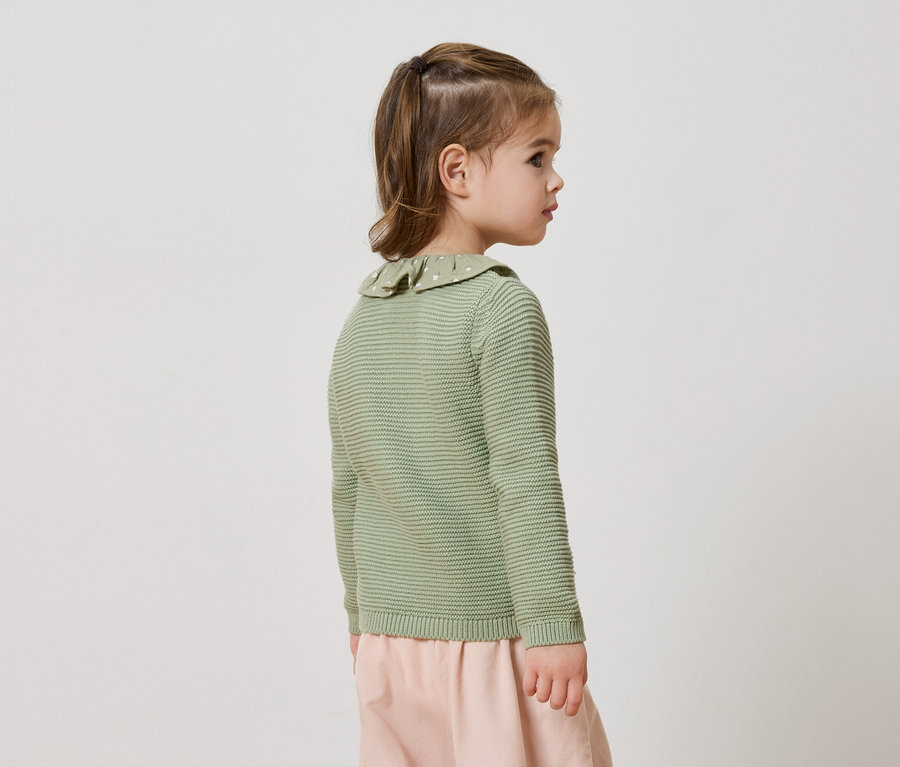 Bočný pohľad na malé dievčatko s chvostom, oblečené v limetkovo zelenom pletenom svetri s volánovým golierom a ružových nohaviciach.