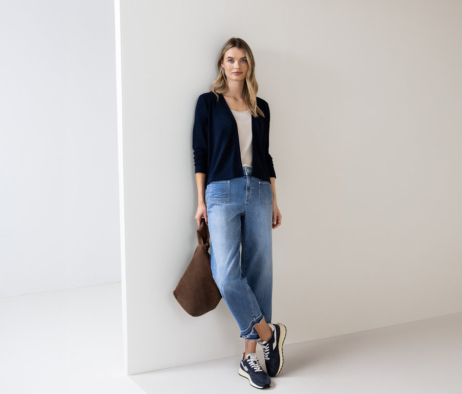 Žena v modrom kardigáne Street One, džínsoch a s hnedou taškou sa opiera o stenu.