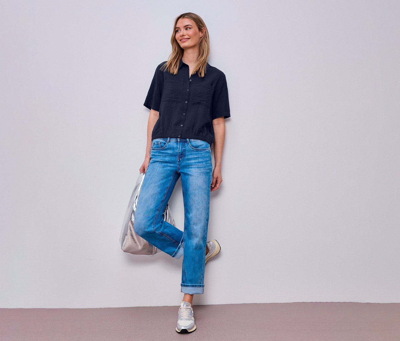 Žena v džínsoch a košeľovej blúzke Street One pózuje so striebornou taškou pri stene.