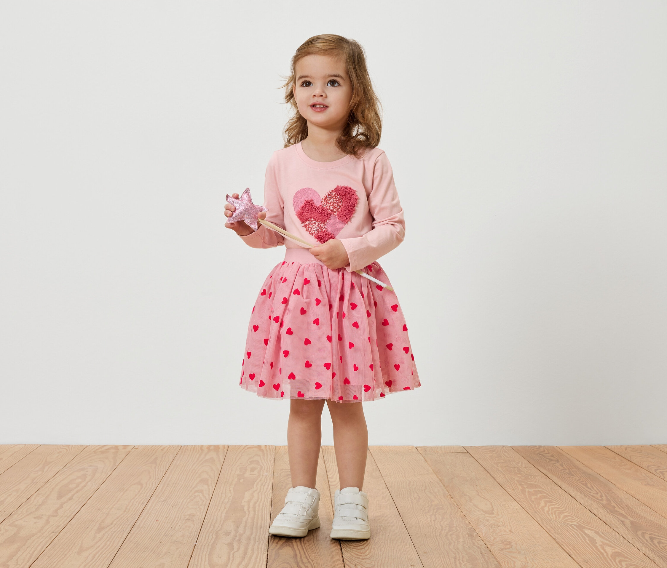 Malé blondínka má na sebe ružové šaty so srdiečkami a drží prútik.