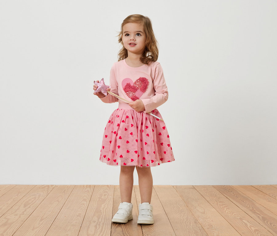 Malé blondínka má na sebe ružové šaty so srdiečkami a drží prútik.