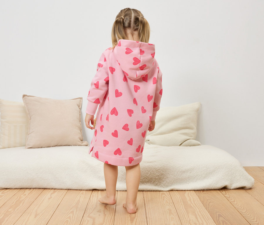 Dievčatko stojí chrbtom k fotoaparátu a má na sebe ružové šaty s kapucňou a srdiečkami.