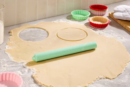 Na pomúčenej pracovnej ploche leží rozvaľkané cesto s dvoma vykrojenými kruhmi, zelený valček na cesto a farebné formičky na tartaletky.